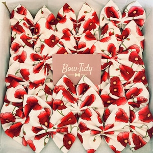 Puede incluir: Una colección de 15 lazos para el pelo de tela blanca con estampado floral de amapolas rojas. Los lazos están dispuestos en una caja y el texto "Bow Tidy" está impreso en una tarjeta rosa en el centro de la imagen.