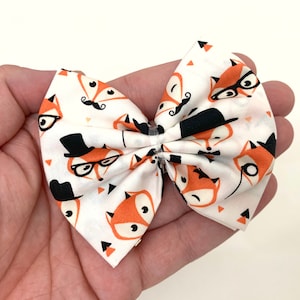 May include: A white fabric bow with a fox print. The foxes are wearing glasses, hats, and mustaches. The bow is tied with a clear elastic band.