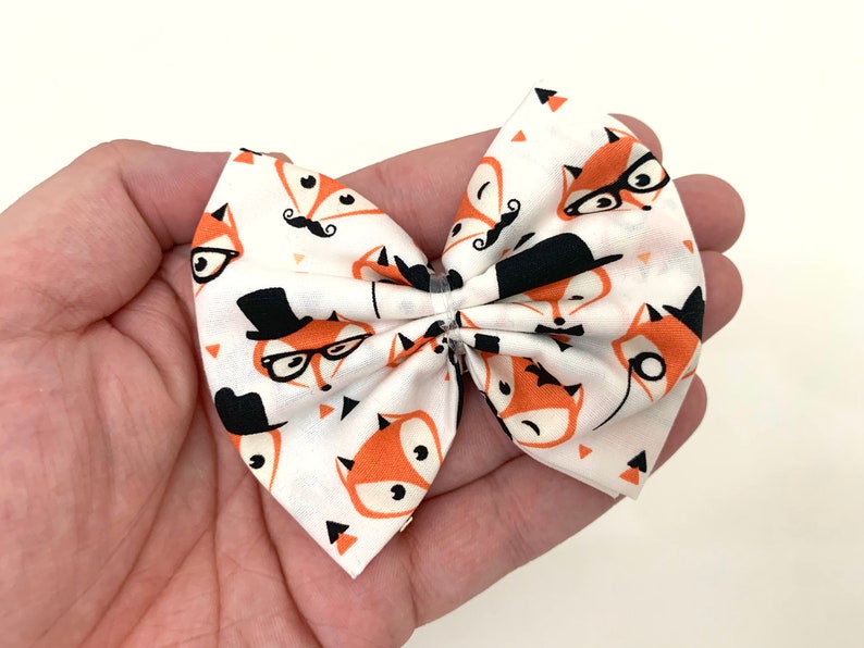 May include: A white fabric bow with a fox print. The foxes are wearing glasses, hats, and mustaches. The bow is tied with a clear elastic band.