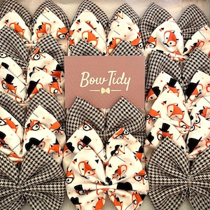 May include: A collection of brown and white bow ties with a fox and top hat pattern. The bow ties are arranged in a grid pattern and have a brown and white houndstooth pattern on the back. The text "Bow Tidy" is printed on a pink card in the center of the image.