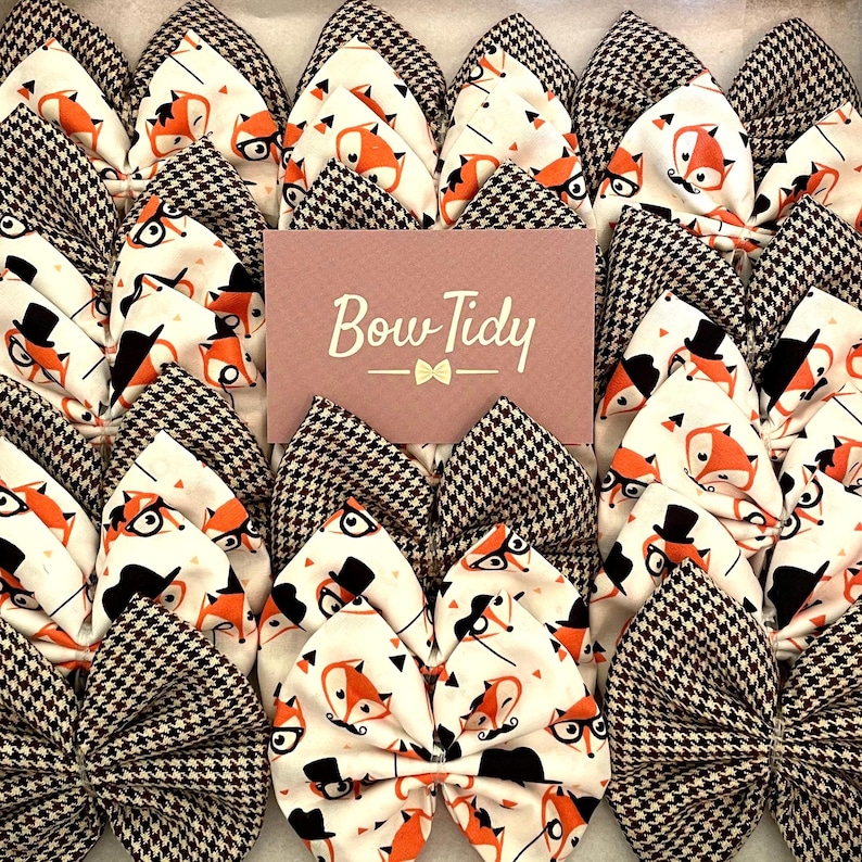 May include: A collection of brown and white bow ties with a fox and top hat pattern. The bow ties are arranged in a grid pattern and have a brown and white houndstooth pattern on the back. The text "Bow Tidy" is printed on a pink card in the center of the image.