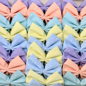 Puede incluir: Colección de lazos de tela de colores pastel con lunares blancos. Los lazos están dispuestos en filas, mostrando una variedad de colores que incluyen azul claro, amarillo, melocotón y lavanda. Los lazos están atados en el centro.
