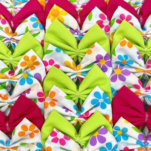 Puede incluir: Una colección de lazos de tela hechos a mano con fondo blanco y estampados florales coloridos. Los lazos están disponibles en tres colores: rosa, verde y blanco.