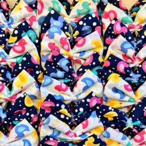 Puede incluir: Primer plano de numerosos lazos de tela, cada uno adornado con patrones de pájaros. Los lazos muestran una gama de colores, como blanco, azul marino, amarillo, rosa y turquesa. Los diseños de pájaros incluyen patos y otras aves, con lunares.