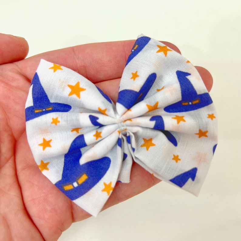 Peut inclure: N&oelig;ud papillon en tissu blanc orn&eacute; de motifs de chapeaux de sorci&egrave;res bleus et d'&eacute;toiles orange. Le n&oelig;ud est nou&eacute; au centre. Il est tenu dans une main.