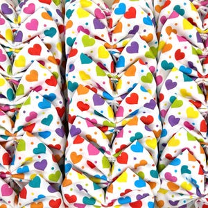 Puede incluir: Primer plano de una tela con un patrón repetido de corazones coloridos sobre un fondo blanco. Los corazones son de varios colores, incluyendo rojo, naranja, amarillo, verde, azul y morado. La tela está doblada y cosida.