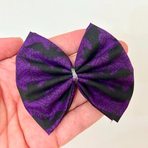 Peut inclure: Un n&oelig;ud papillon en tissu fait &agrave; la main avec un motif sur le th&egrave;me d'Halloween. Le n&oelig;ud est principalement violet avec un motif de toile d'araign&eacute;e noire. Il est nou&eacute; au centre avec du fil argent&eacute;.