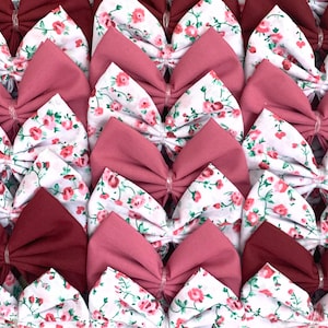May include: A collection of handmade fabric bows in a variety of floral patterns. The bows are made with a combination of white fabric with pink floral print and solid burgundy fabric.
