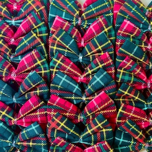 Peut inclure: Une collection de nœuds en tissu disposés en grille, chacun présentant un motif écossais vibrant avec des lignes entrecroisées de vert foncé, de rouge vif et de fines rayures jaunes. Chaque nœud est fixé au centre par une attache transparente.