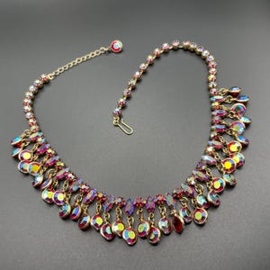 May include: A vintage necklace featuring iridescent, teardrop-shaped pendants and round, colourful gemstones. The necklace has a gold-toned chain and clasp. The gemstones display a range of colours, including red, purple, and yellow, creating a dazzling effect.