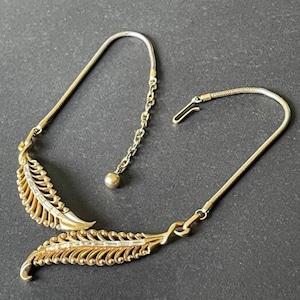 Op de afbeelding: Een goudkleurige ketting met een bladvorm. De ketting heeft twee gestileerde bladhangers met ingewikkelde details, verbonden door een delicate ketting en een slangenketting. Het heeft een haaksluiting.