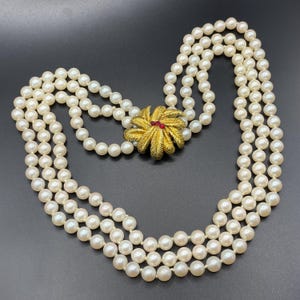Pode incluir: Um colar de pérolas de três fios com um broche de flor dourado e vermelho.