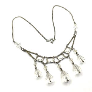 May include: A silver-toned necklace with clear crystal accents. The necklace features a chain, geometric shapes, and dangling teardrop-shaped crystals. The design is symmetrical, with the crystals evenly spaced along the bottom.