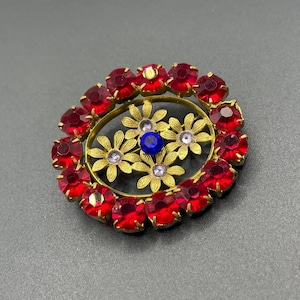 Peut inclure: Broche ovale avec un cadre doré. La broche présente un anneau de pierres précieuses rouges entourant un motif floral. Le centre du motif floral comporte une pierre précieuse bleue, avec de plus petites pierres précieuses dans les fleurs.