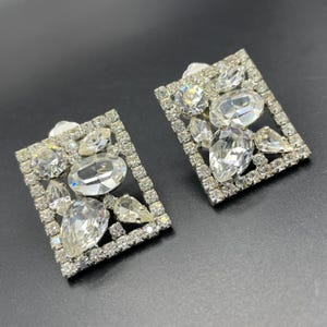 Puede incluir: Un par de pendientes de clip de tono plateado con un diseño rectangular. Los pendientes están adornados con un grupo de diamantes de imitación transparentes de varios tamaños y formas.