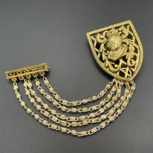 May include: A gold-tone metal brooch featuring a shield-shaped crest with an embossed helmet design. The crest is connected by five draped, textured link chains to a rectangular bar pin, creating a layered, multi-strand chain effect.