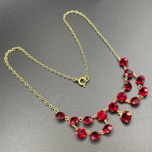 May include: A gold-toned necklace with a series of vibrant red gemstones. The necklace features a delicate chain and a cluster of round, ruby-colored stones. The gemstones are set in a gold-colored metal, creating a striking contrast.