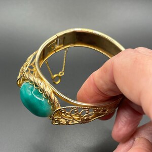 以下が含まれることがあります： 金色のヒンジ付きバングルブレスレット。楕円形のターコイズ色の石が特徴です。ブレスレットは、複雑なフィリグリーと石の周りの装飾的なロープのようなボーダーが特徴です。留め金が見え、ブレスレットは暗い背景に対して保持されています。