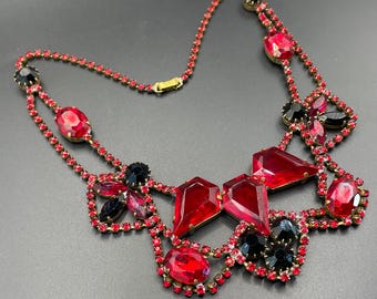 May include: A vintage necklace featuring a gold-tone chain and setting, adorned with vibrant red and black rhinestones. The necklace showcases a bib-style design with various shapes and sizes of stones, creating a bold and elegant statement piece.