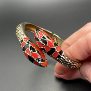 Könnte beinhalten: Ein goldfarbenes Metallarmband in Form einer Schlange. Der Schlangenkopf ist rot und schwarz, mit einem schuppenartigen Muster. Das Armband wird in einer Hand vor dunklem Hintergrund gehalten.