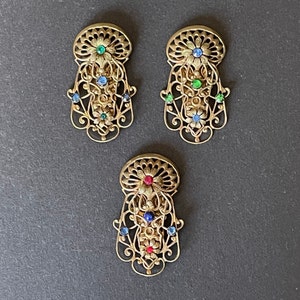 Peut inclure: Trois pinces à cheveux en filigrane dorées avec des strass colorés. Chaque pince présente une combinaison de couleurs différente de strass bleus, verts et rouges.