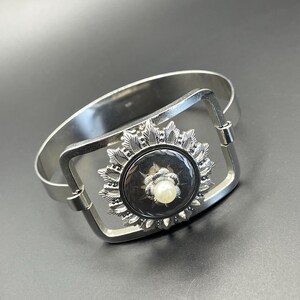 Puede incluir: Brazalete plateado con un centro rectangular. El centro presenta un diseño circular negro con un borde floral plateado y una perla central. El brazalete tiene un cierre de bisagra.
