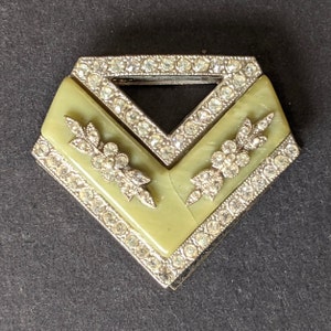May include: A silver and green geometric brooch with a V-shaped design. The brooch is decorated with rhinestones and has a floral motif.