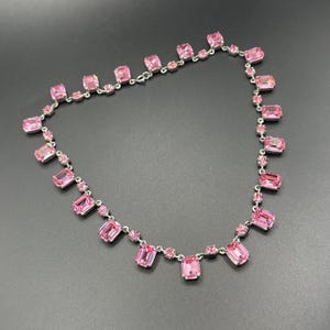 May include: A silver necklace with pink rectangular and round gemstones. The necklace has a delicate chain. The gemstones are a vibrant pink colour and are set against a dark background.