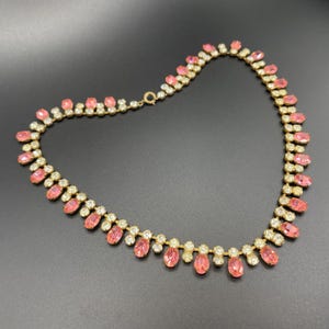 May include: A vintage necklace with a gold-tone metal chain. The necklace features alternating oval pink and round clear gemstones. The pink gemstones are set in a teardrop pattern, while the clear gemstones are set in a row. The necklace is displayed on a black surface.