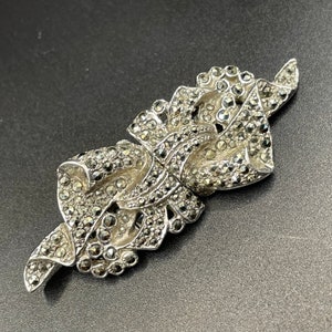 Puede incluir: Un broche de plata con un diseño floral hecho de piedras de marcasita. El broche tiene la forma de dos hojas superpuestas con detalles intrincados.