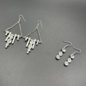 Può includere: Orecchini color argento con cristalli trasparenti. Sono mostrate due paia. Un paio presenta un design geometrico con catena e più cristalli. L'altro paio ha un design lineare semplice con tre cristalli.