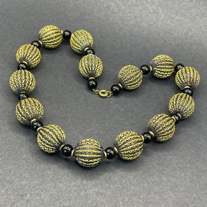 Puede incluir: Un collar de oro con cuentas negras y un diseño texturizado. El collar está hecho de cuentas redondas con un patrón tejido.