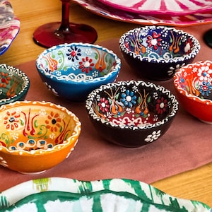 May include: Six small decorative bowls with colourful floral patterns. The bowls are made of ceramic and have a white rim. The bowls are arranged on a pink and brown table runner.