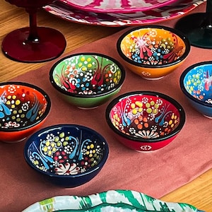 May include: Six small colourful ceramic bowls with intricate floral patterns. The bowls are arranged on a pink table runner with a wooden surface underneath.