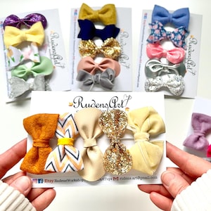 May include: Several sets of handmade hair bows in various colors and patterns. Each set includes multiple bows, with colors ranging from glittery gold and silver to solid hues of yellow, blue, pink, and green. The bows are displayed on white cards with the text "Rudens Art".