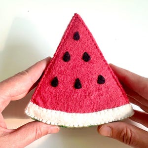 May include: A triangular watermelon slice crafted from felt, featuring a bright pink interior with dark brown embroidered seeds, a white rind section, and visible white stitching along the edges of the fabric construction.