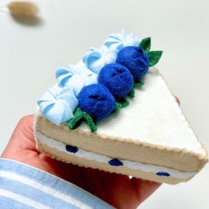 Può includere: Una fetta di torta in feltro con glassa bianca, riccioli di glassa azzurra e mirtilli in feltro blu scuro. La torta ha una base beige e un ripieno bianco con accenti blu. Sono presenti anche foglie di feltro verdi.