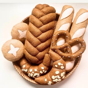 Peut inclure: Un panier tressé rempli de divers pains en feutrine, comprenant une grosse miche tressée, deux petits pains ronds avec des centres en forme d'étoile, deux baguettes, un bretzel à points blancs et deux croissants garnis de flocons blancs.