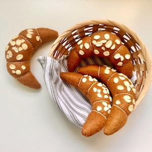 Op de afbeelding: Vier halvemaanvormige viltgebakjes in een bruine tint, versierd met kleine witte, amandelvormige viltstukjes. Drie van de gebakjes liggen in een ronde mand van gevlochten riet, bekleed met een wit en grijs gestreepte stoffen doek.