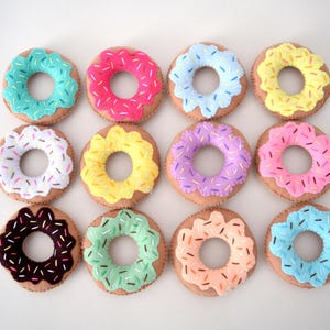 May include: A collection of twelve circular felt donut toys arranged in a grid. Each donut features a tan base with a colorful frosted top in shades of teal, pink, blue, yellow, white, purple, brown, mint, and peach, decorated with small sprinkles.