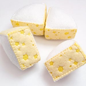 Puede incluir: Cuatro cuñas de queso de fieltro con tapas blancas y lados amarillos perforados. Las cuñas de queso están dispuestas sobre una superficie blanca. El queso de fieltro es un juguete o un artículo decorativo.