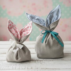 Peut inclure: Deux sacs cadeaux en tissu gris en forme de lapins de Pâques. L'un a des oreilles à motifs géométriques roses et blancs, noué avec un nœud en dentelle blanche. L'autre a des oreilles rayées bleues et blanches, noué avec un ruban à pois turquoise.
