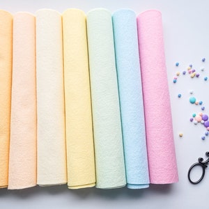 Puede incluir: Siete rollos de fieltro de color pastel en tonos melocotón, naranja claro, amarillo, verde claro, azul claro y rosa. El fieltro está enrollado y los extremos son visibles. También hay cuentas de colores y un par de tijeras negras sobre una superficie blanca.