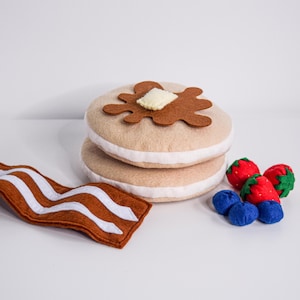 May include: A felt breakfast set featuring two pancakes with butter and syrup, a strip of bacon, and berries. The pancakes are beige with white filling. The bacon is brown with white stripes. The berries are red and blue.