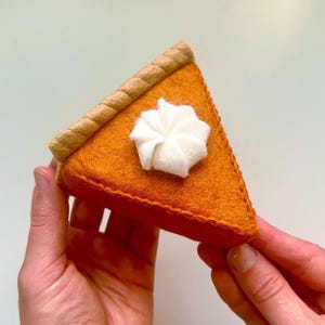 Puede incluir: Una porción triangular de tarta de calabaza hecha de fieltro naranja, con una corteza trenzada de color tostado en el borde posterior y un copete de nata montada de fieltro blanco en el centro. Los bordes están cosidos con hilo naranja.