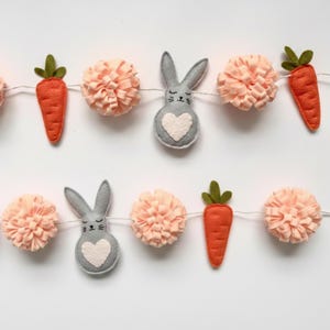 以下が含まれることがあります： フェルトのニンジン、ピーチ色のポンポン、ハート型の腹を持つ灰色のウサギのフィギュアが特徴の、お祝いのイースターガーランド。 ガーランドは細い白い紐でつながれており、春の装飾に最適です。