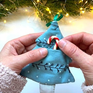 Könnte beinhalten: Ein hellblauer Weihnachtsbaumschmuck aus Stoff mit einer kleinen Tasche, die eine rot-weiß gestreifte Miniatur-Zuckerstange enthält. Der Baum ist mit schwarzen Zweigillustrationen und weißen Punkten verziert und hat eine grüne Bandschlaufe.