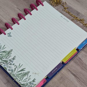 May include: A spiral-bound planner with lined pages and pink plastic rings. The planner features botanical illustrations and month tabs. The words "My Notes" are printed at the top of the page. A gold-colored bookmark is also present.
