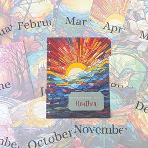 May include: A planner with a stained-glass style cover featuring a vibrant sunrise over ocean waves in shades of red, orange, yellow, and blue. A central label reads "THIS PLANNER BELONGS TO Heather." The planner is surrounded by monthly calendar pages.