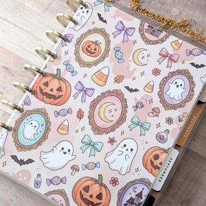 May include: A disc-bound planner featuring a light pink cover with a repeating pattern of orange jack-o'-lanterns, white ghosts, candy corn, pastel bows, crescent moons, and ornate frames. The planner includes tabbed dividers labeled September and December.